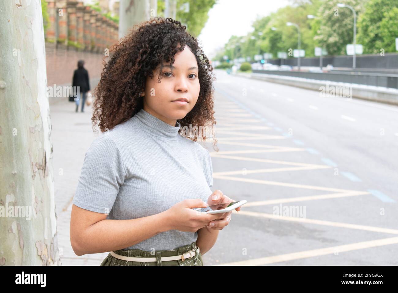 giovane ragazza nera con capelli ricci in città in attesa di un taxi o uber con cellulare in mano. Foto orizzontale. Foto Stock
