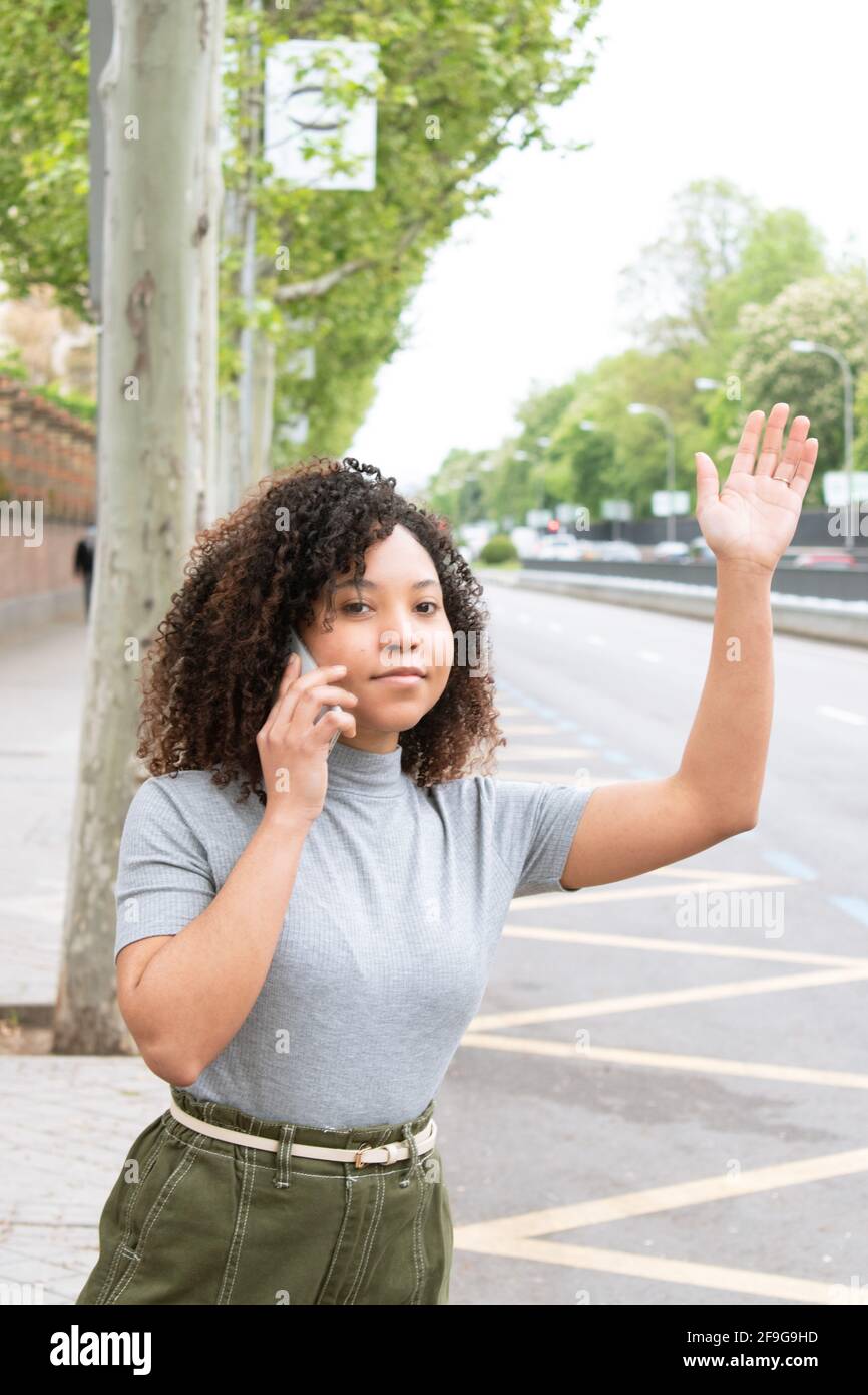 Giovane ragazza nera con capelli ricci alza la mano per chiamare un uber mentre parla sul suo cellulare in città. Fotografia verticale. Foto Stock
