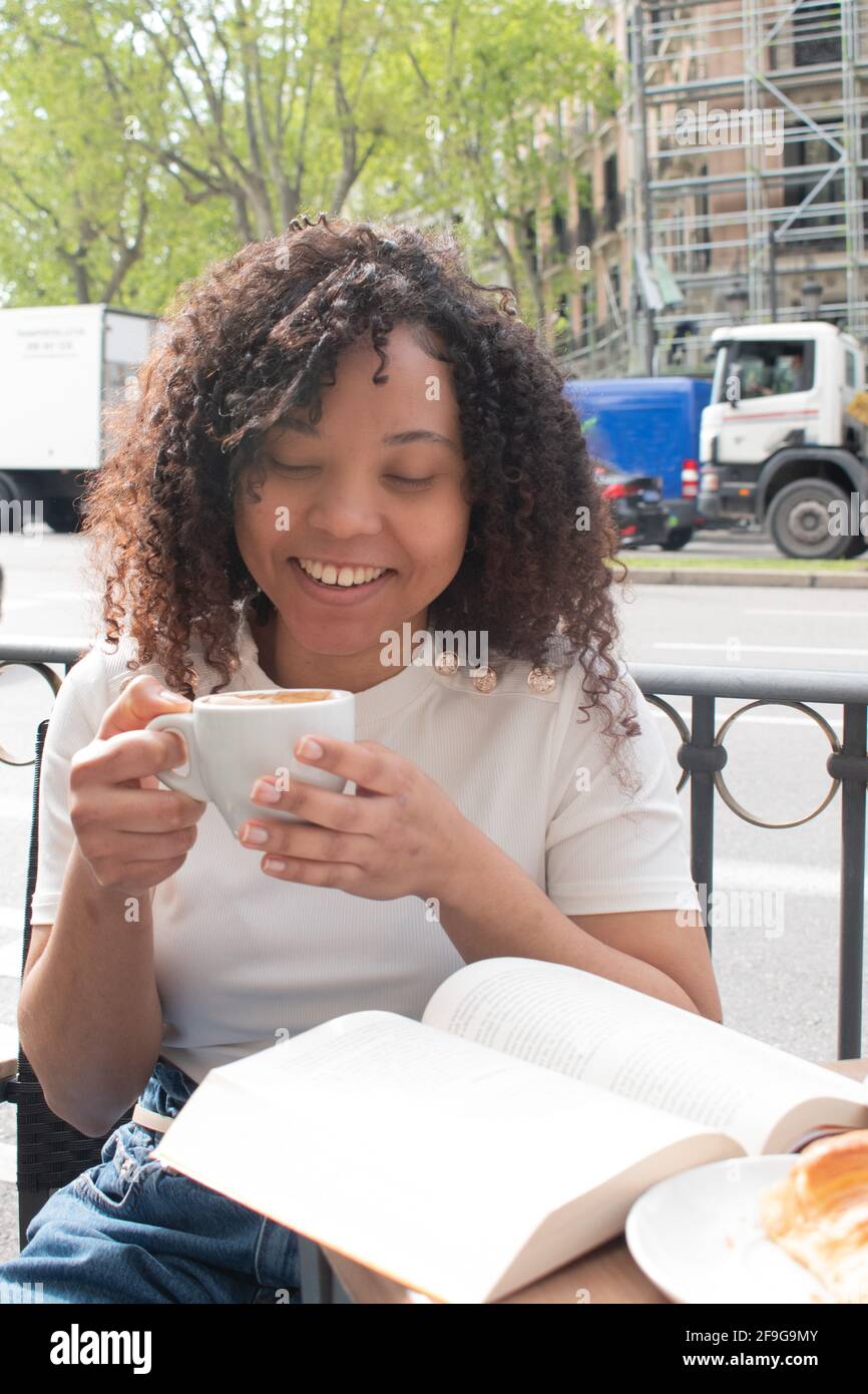 giovane donna nera con ricci che beve un caffè e che legge un libro all'esterno di una caffetteria. Foto Stock