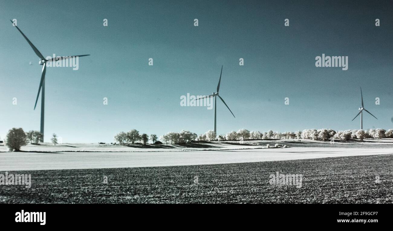 Una fila di tre turbine eoliche che torreggiano nel cielo sopra alberi e campi infrarossi Foto Stock