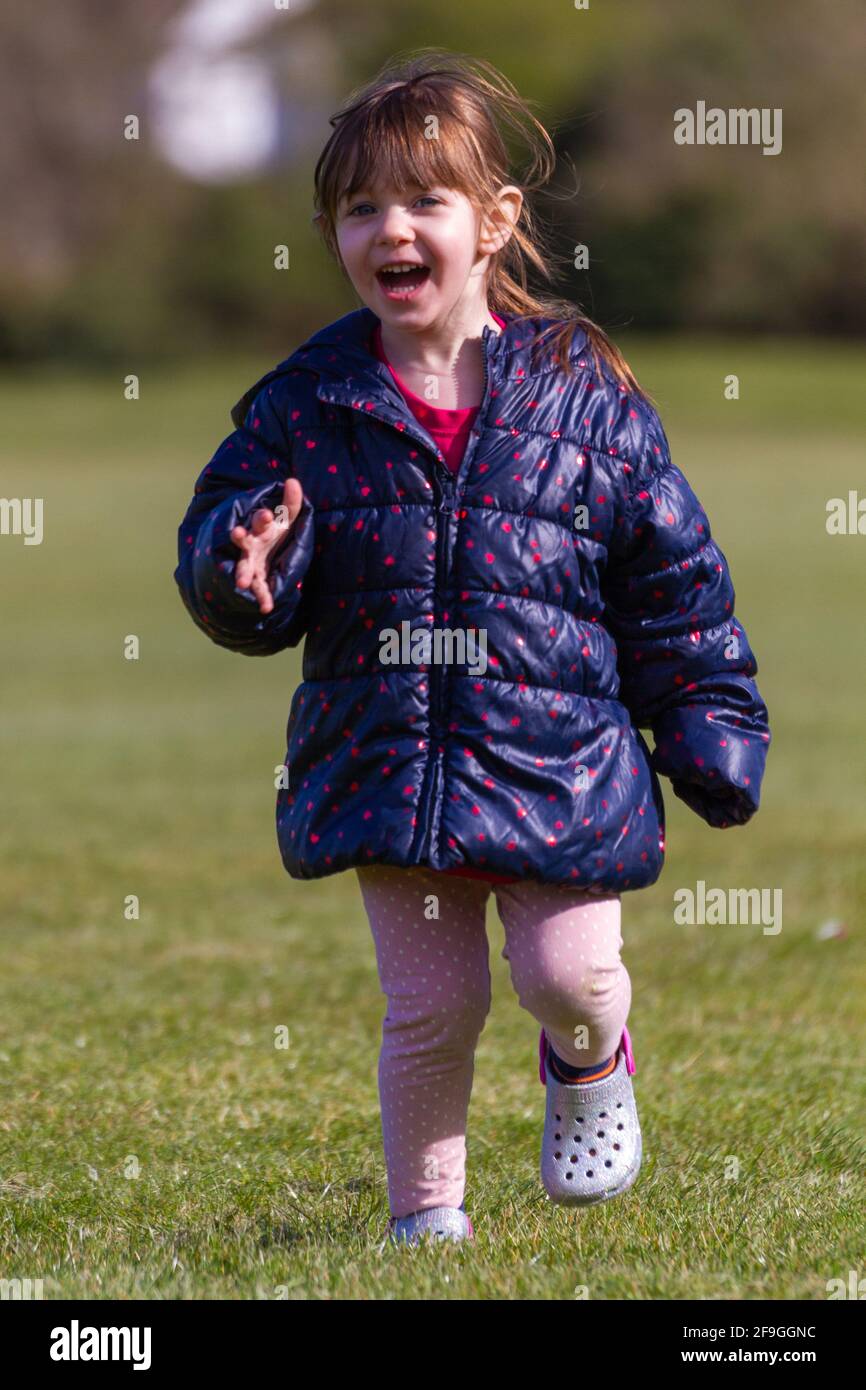 Foto a tutta lunghezza di una bambina carina marrone con capelli una giacca blu e pantaloni rosa che corrono sull'erba in un parco in una soleggiata giornata di primavera Foto Stock