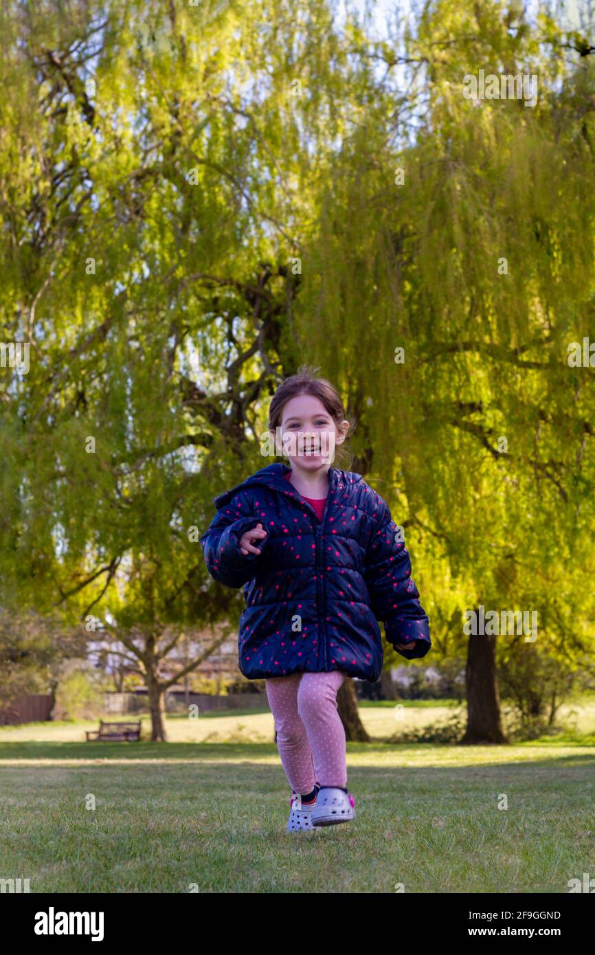 Foto a tutta lunghezza di una bambina carina marrone con capelli una giacca blu e pantaloni rosa che corrono sull'erba in un parco in una soleggiata giornata di primavera Foto Stock