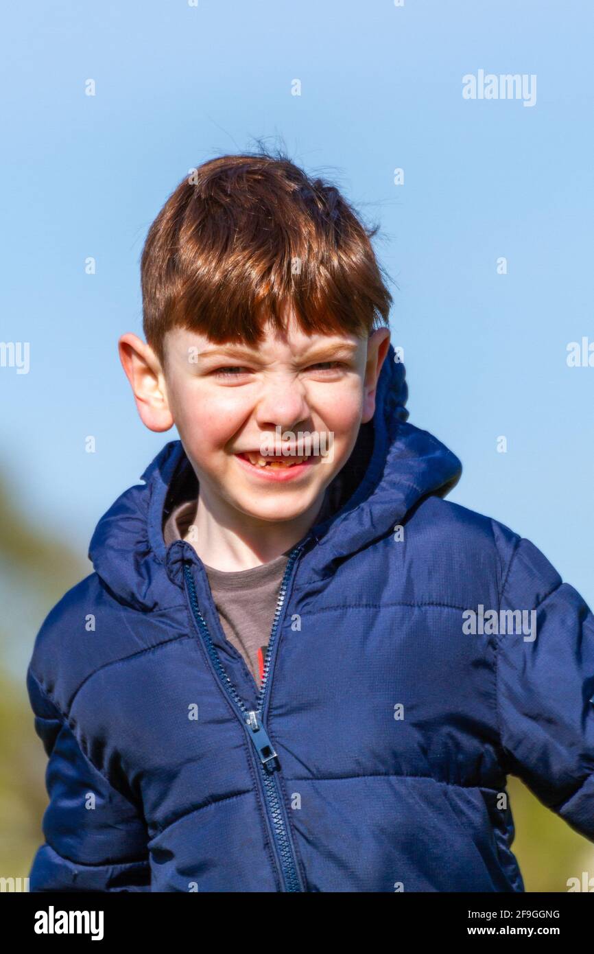 Un simpatico ragazzo rosso dagli occhi azzurri che indossa una giacca blu che corre in un parco in una soleggiata giornata primaverile Foto Stock