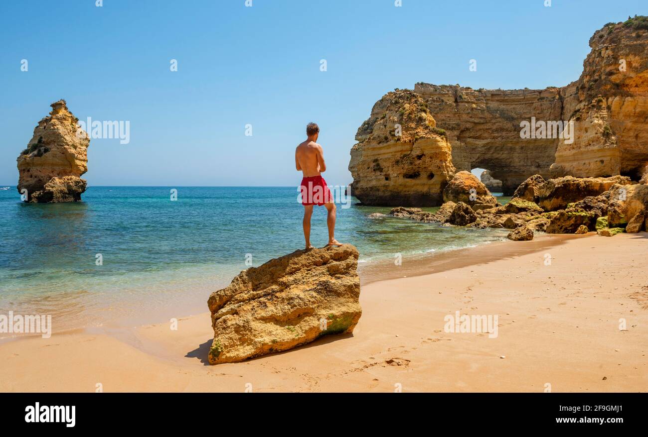 Giovane uomo in piedi su una roccia, Praia da Marinha spiaggia, costa rocciosa, formazione rocciosa in mare, Algarve, Lagos, Portuga Foto Stock