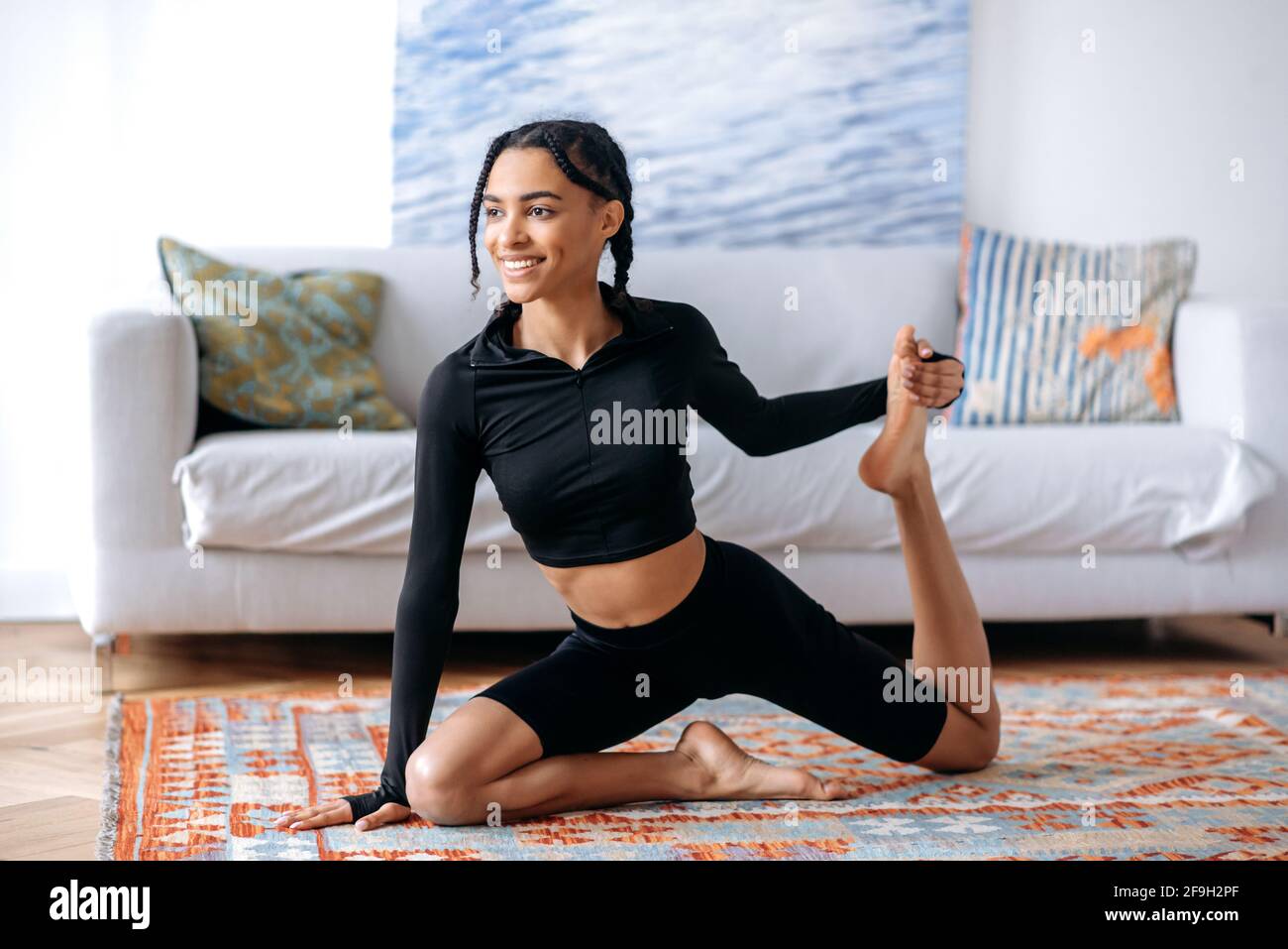 Flessibile giovane bella donna afroamericana in abbigliamento sportivo, è impegnata fitness, conduce uno stile di vita sano, si allunga a casa su un pavimento, sorride, sembra felice Foto Stock