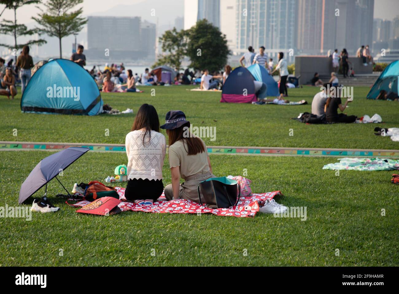 20 3 2021: Due donne cinesi, famiglie di amici che si siedono e si rilassano sul campo di erba a Tamar Park, Hong Kong durante Covid-19, con molti altri simili Foto Stock