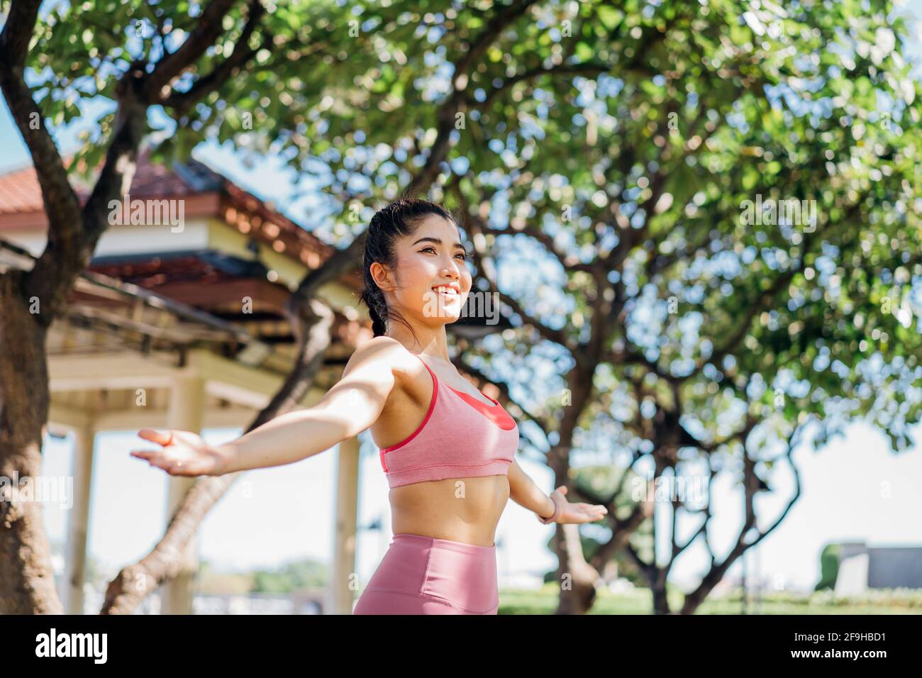 Attraente e misura giovane donna asiatica sorridente che pratica yoga con braccia stirate in un parco all'aperto durante la routine mattutina per un corpo sano Foto Stock