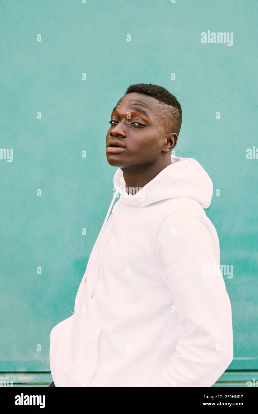 Giovane uomo nero africano contro un muro verde guardando fotocamera che indossa una felpa bianca con stile e sicurezza Foto Stock