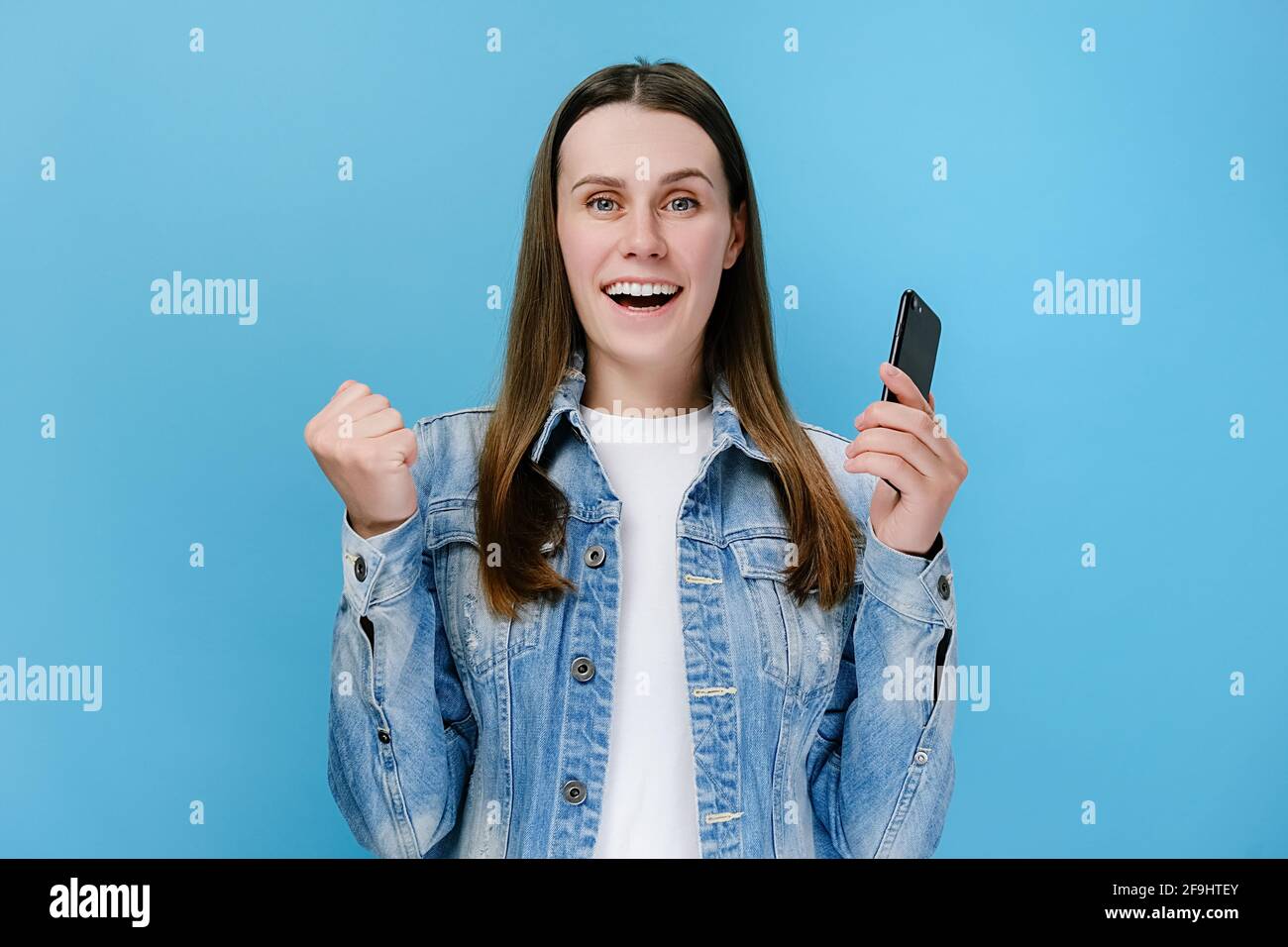 Ecited giovane donna tiene smartphone ricevere sms con grandi notizie si sente felice fare sì gesto, indossa una giacca denim, isolato su sfondo blu Foto Stock