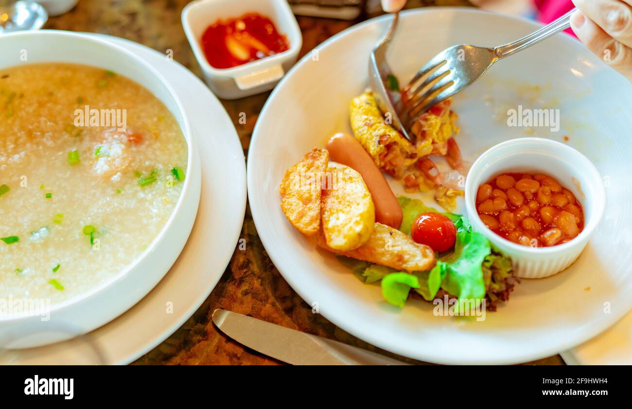 La persona usa forchetta e cucchiaio per la colazione al ristorante. Porridge di riso o gruel di riso con gamberi in ciotola bianca e fette di salsiccia e patate fritte. Foto Stock