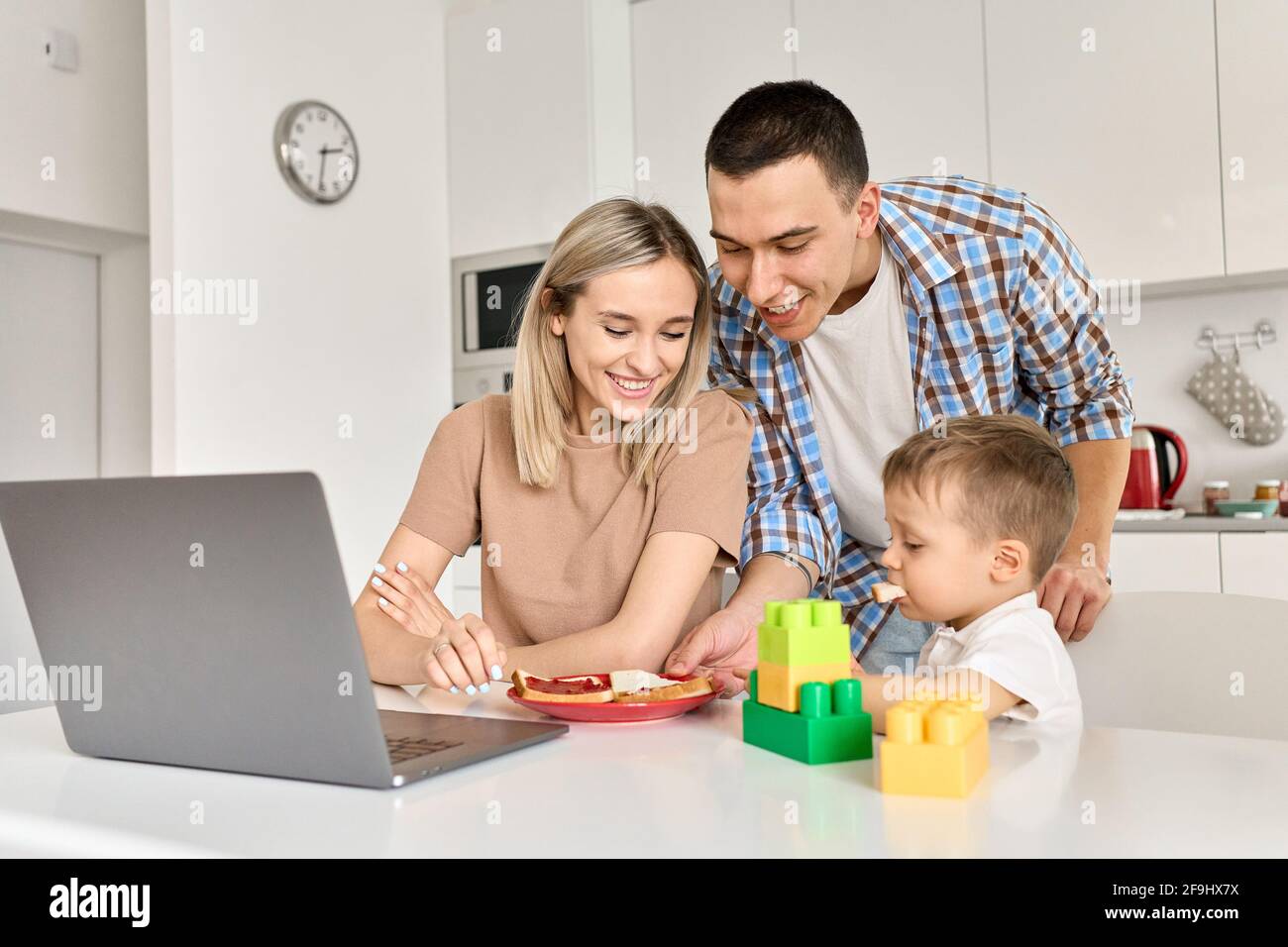 Felice famiglia giovane con figlio di capretto che mangia toast usando il laptop in cucina. Foto Stock