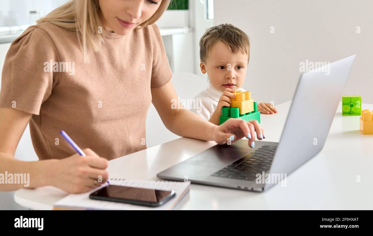 Madre impegnata che lavora a casa utilizzando il computer portatile mentre il bambino gioca. Foto Stock