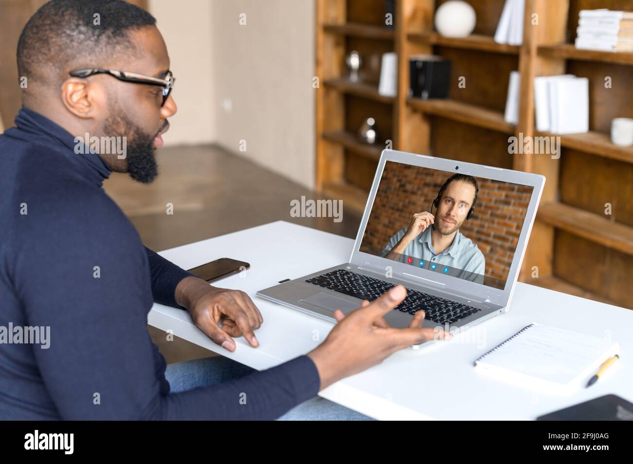 Uomo d'affari afro-americano si sente felice di aver chiamato con un collega maschio in cuffia utilizzando un'applicazione speciale, parlare di fronte alla webcam, briefing online o riunione, impegnato in conferenza web con il partner Foto Stock