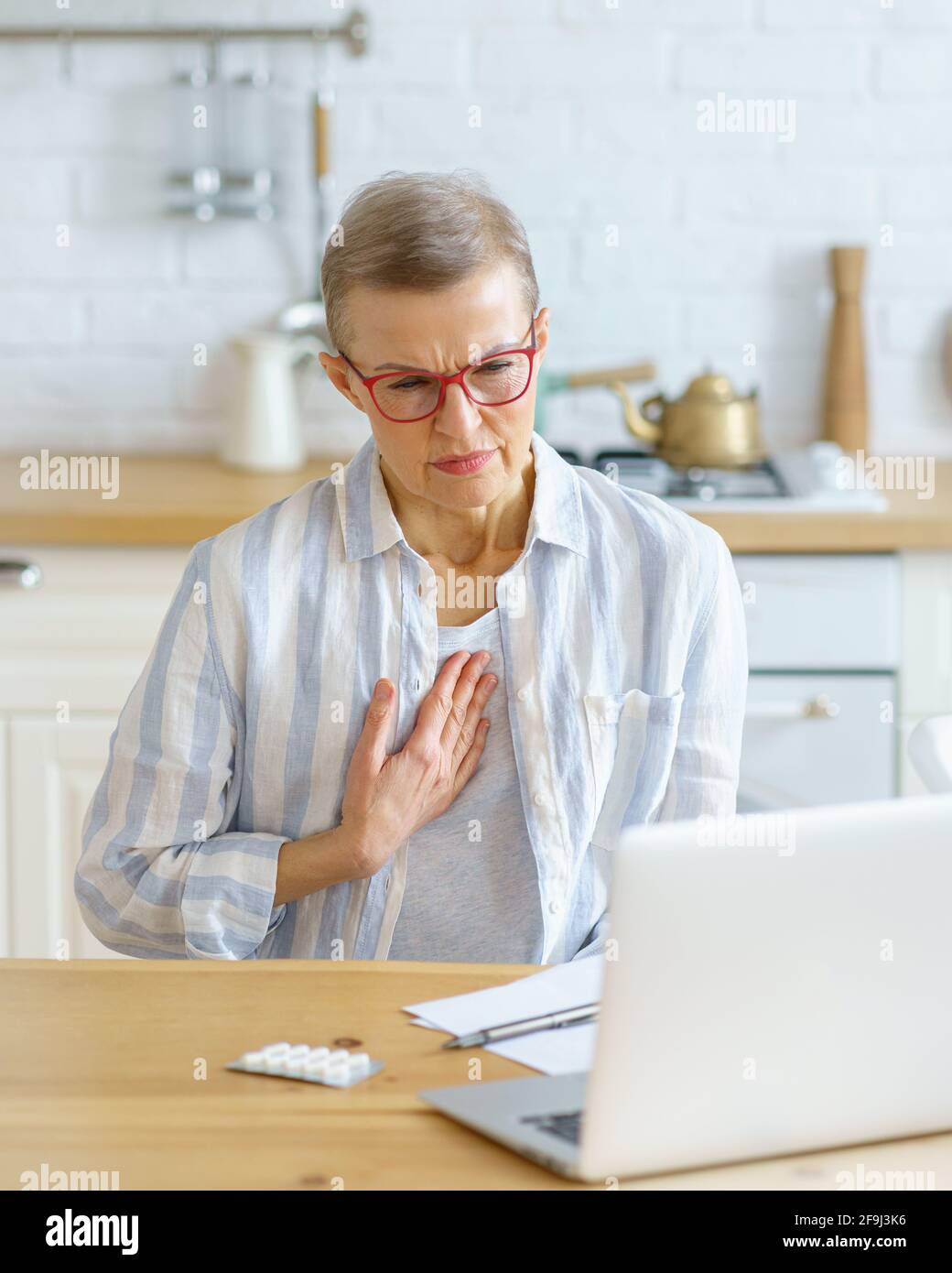 Triste malato donna di mezza età che soffre di dolore al petto o tosse avendo consultazione online con il medico sul computer portatile durante l'epidemia di covid 19, seduto in cucina a casa. Concetto di sanità e medicina Foto Stock