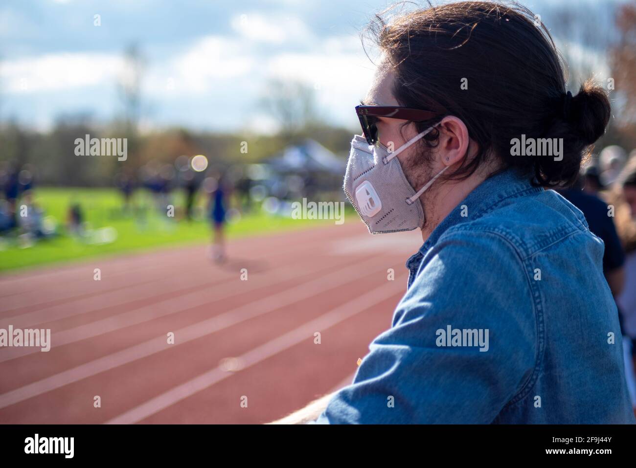 Un giovane uomo caucasico in una camicia blu in denim indossa occhiali da sole e una maschera facciale mentre guarda una pista e un evento atletico di campo ant una pista da runnning. Chiudi Foto Stock