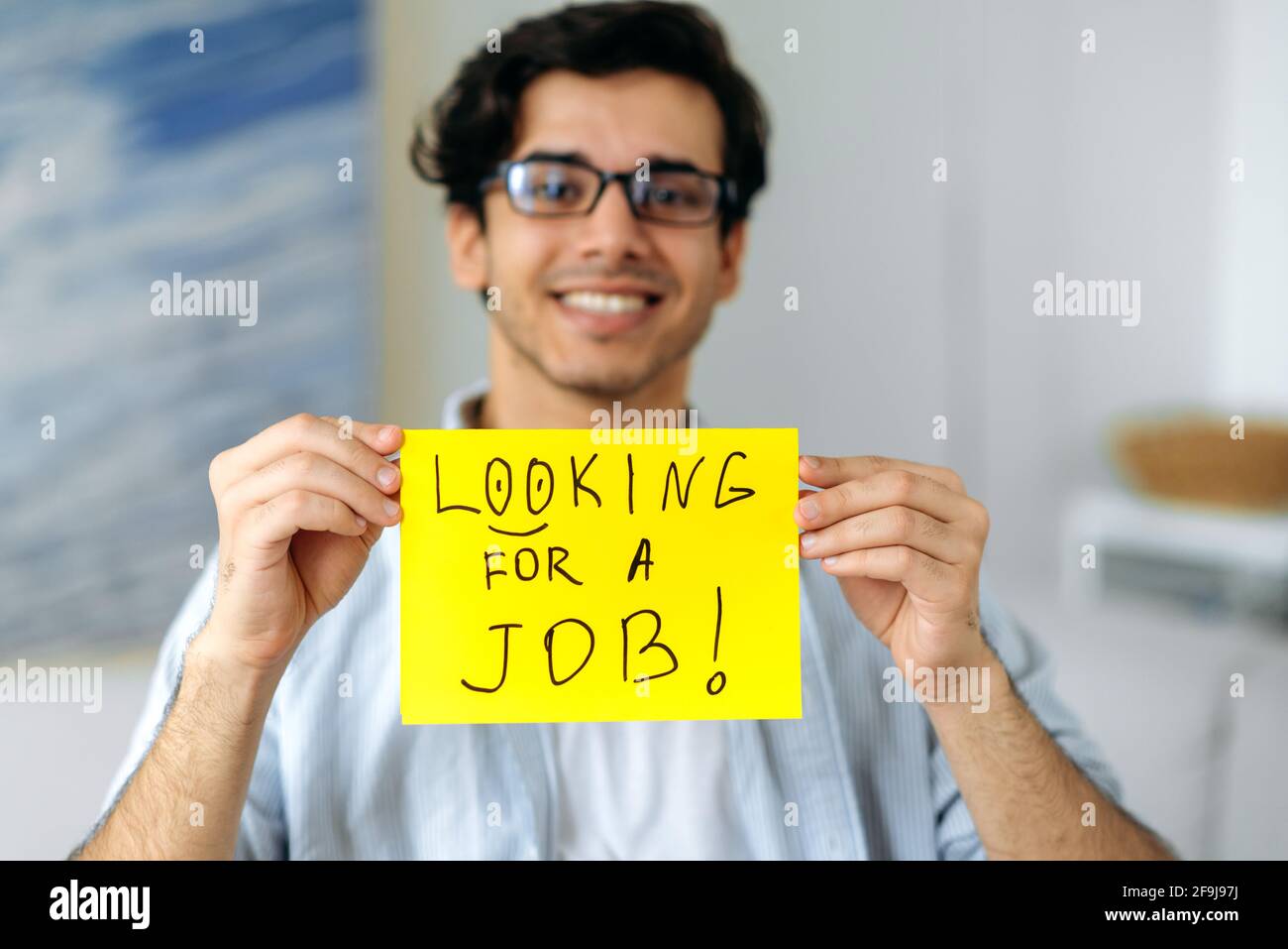 Defocused simpatico ragazzo di corsa misto intelligente con occhiali, studente o disoccupato, tenendo un segno con l'iscrizione alla ricerca di un lavoro, guardando la macchina fotografica, sorridente amichevole Foto Stock