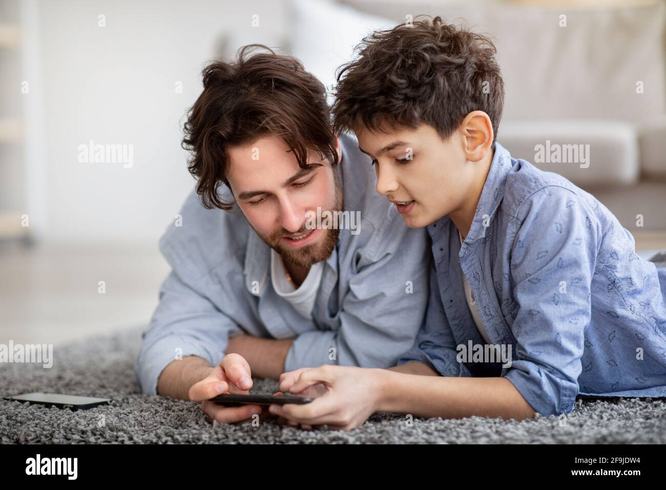 Padre e figlio usano lo smartphone, guardando le foto o il ragazzo che aiuta il papà con il gioco, trascorrendo il tempo a casa Foto Stock