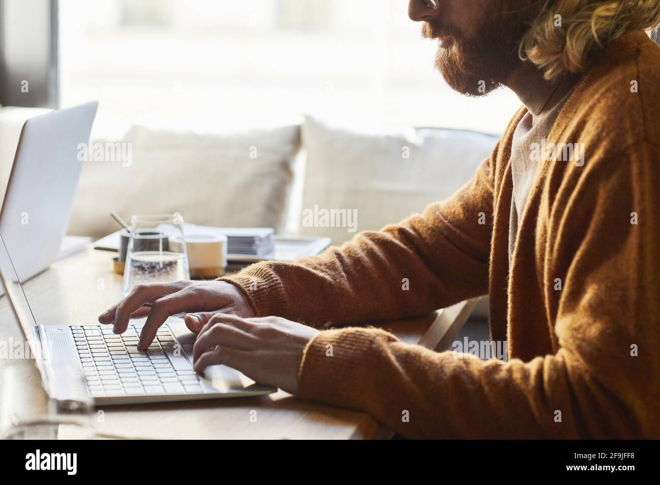 Minimo ritaglio di uomo bearded utilizzando il computer portatile mentre si lavora al bar tavolo illuminato dalla luce del sole in calde tonalità marrone, spazio di copia Foto Stock