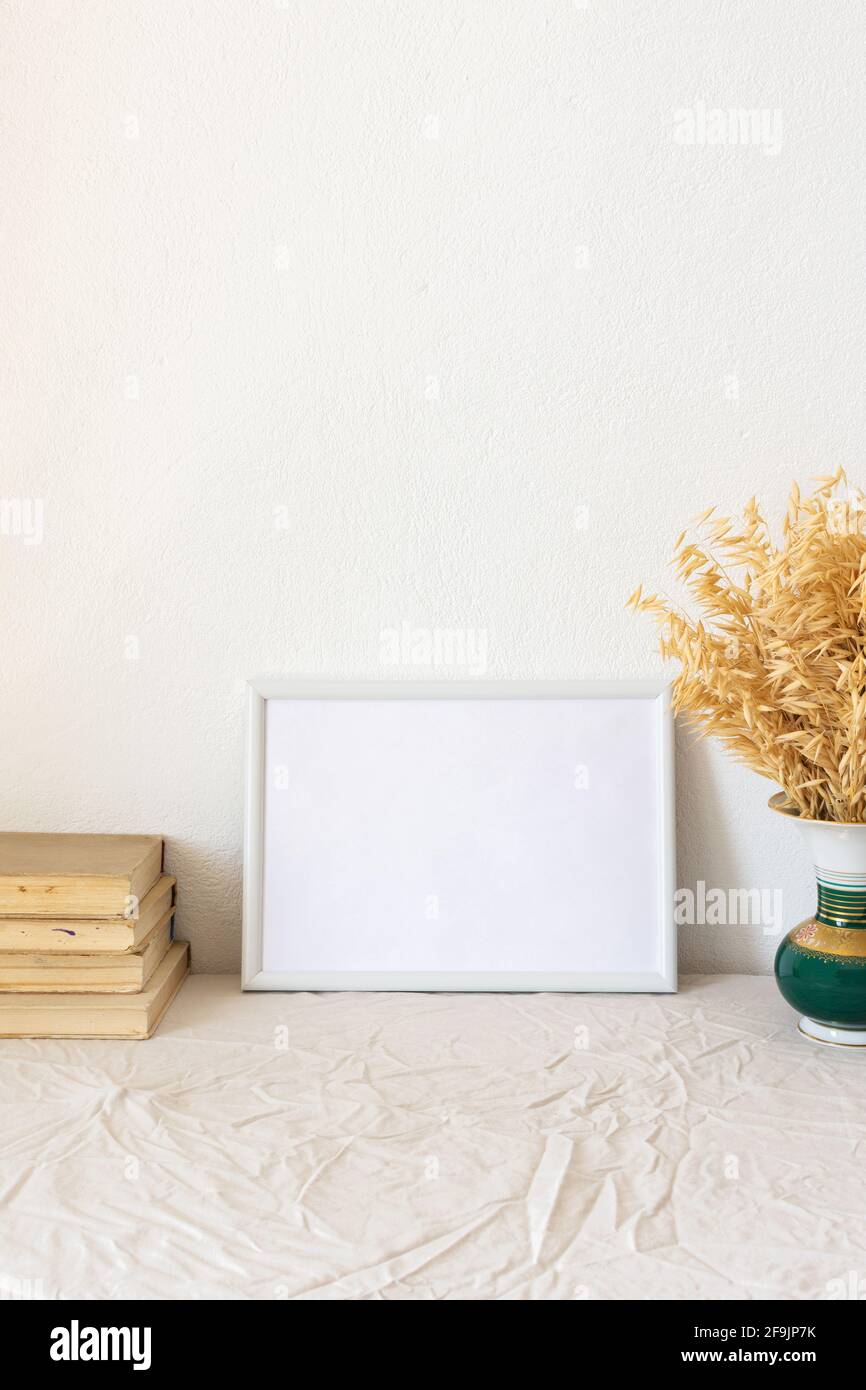 Home decor mock-up, quadro vuoto cornice vicino bianco dipinto muro di cemento, vaso con secchi gambi di avena gialla, vecchi libri Foto Stock