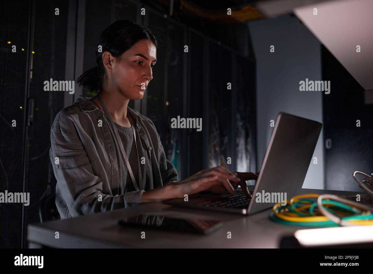Ritratto di un tecnico di rete femminile che utilizza un computer portatile mentre si è seduti in una stanza server buia, copia spazio Foto Stock