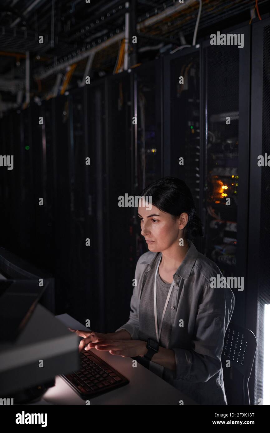 Ritratto verticale di un tecnico di rete femminile che utilizza il computer mentre lavora in una stanza server buia, spazio di copia Foto Stock