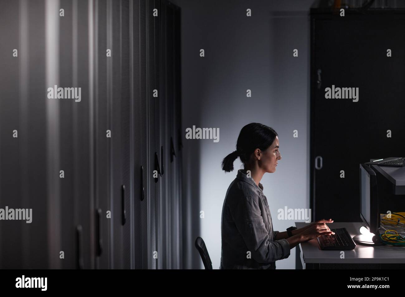 Vista laterale grafica ritratto di un tecnico di rete femminile che utilizza il computer mentre lavora in una stanza server buia, spazio di copia Foto Stock