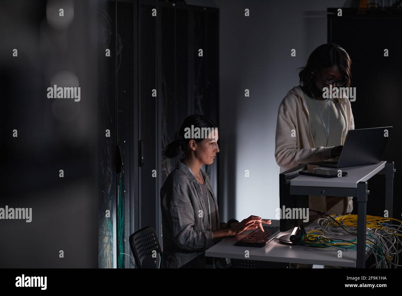 Ritratto di due ingegneri IT femminili che lavorano con il computer in una stanza server buia, spazio di copia Foto Stock