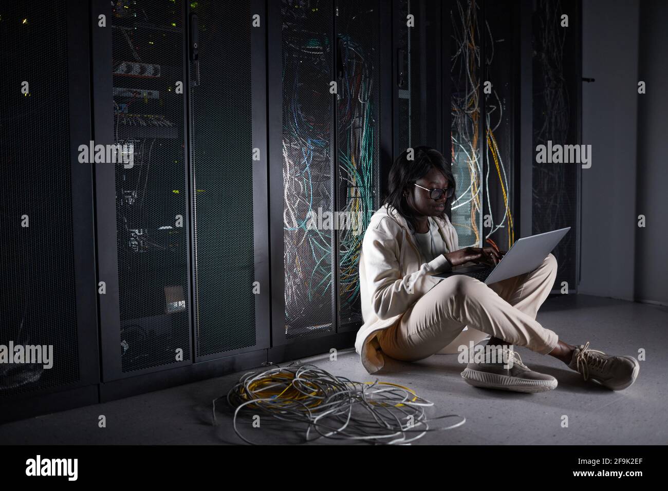 Ritratto di una giovane donna afroamericana seduto sul pavimento nella sala server e utilizzando un computer portatile mentre lavora con il supercomputer nel data center, Foto Stock