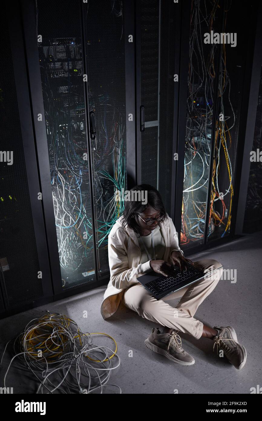 Ritratto verticale a lunghezza intera della giovane afroamericana seduta pavimento nella sala server e utilizzo di un computer portatile mentre si lavora con supercomputer al dat Foto Stock