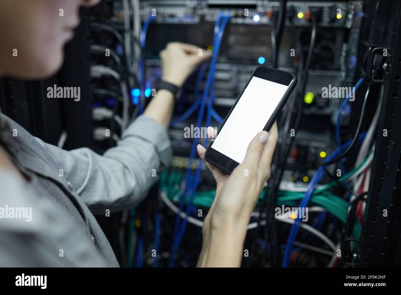 Primo piano di un tecnico di rete femminile che tiene lo smartphone con schermo vuoto mentre si collegano i cavi nel cabinet del server, spazio di copia Foto Stock