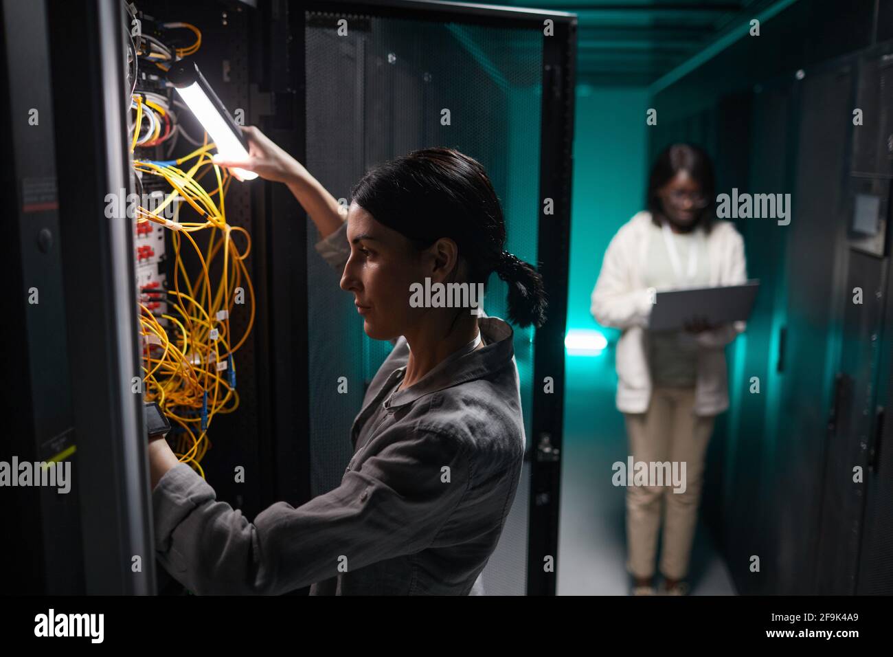 Vista laterale verticale del tecnico di rete femmina che collega i cavi nell'armadietto del server mentre lavora con il supercomputer nel data center, spazio di copia Foto Stock