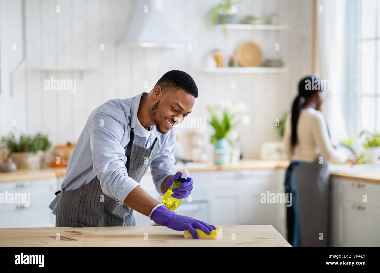 Bel ragazzo nero in guanti di gomma che pulisce tavolo da cucina con  detergente, sua moglie che lava i piatti sullo sfondo Foto stock - Alamy