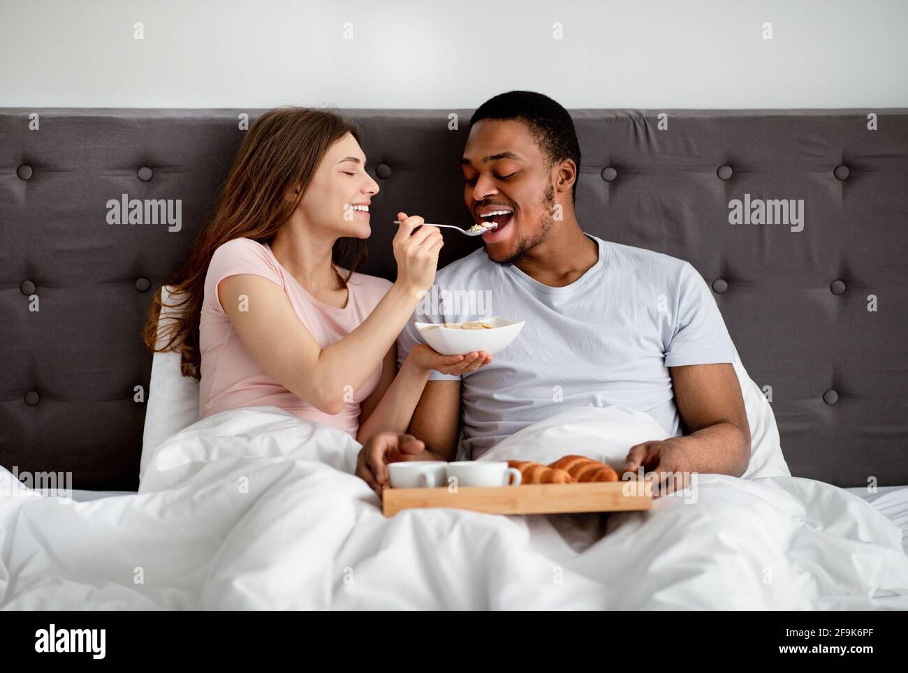 Felice coppia multirazziale che ha fatto colazione a letto, giovane donna che alimenta il marito con gustosi cereali Foto Stock