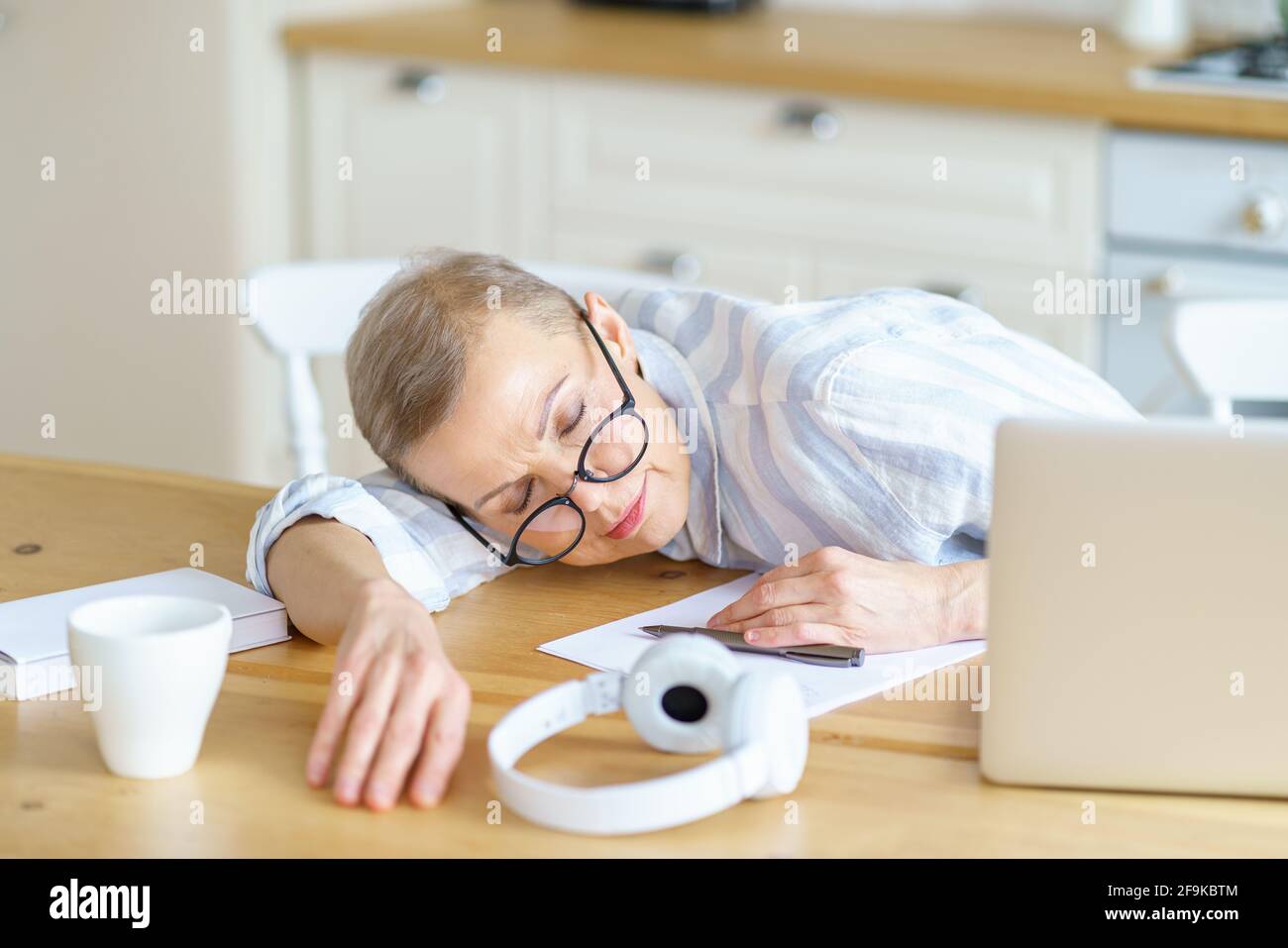 Donna stanca di mezza età che indossa occhiali che dormono su un tavolo di legno in cucina mentre lavora o studia su un computer portatile online da casa. Persone anziane e tecnologie, concetto di lavoro remoto Foto Stock