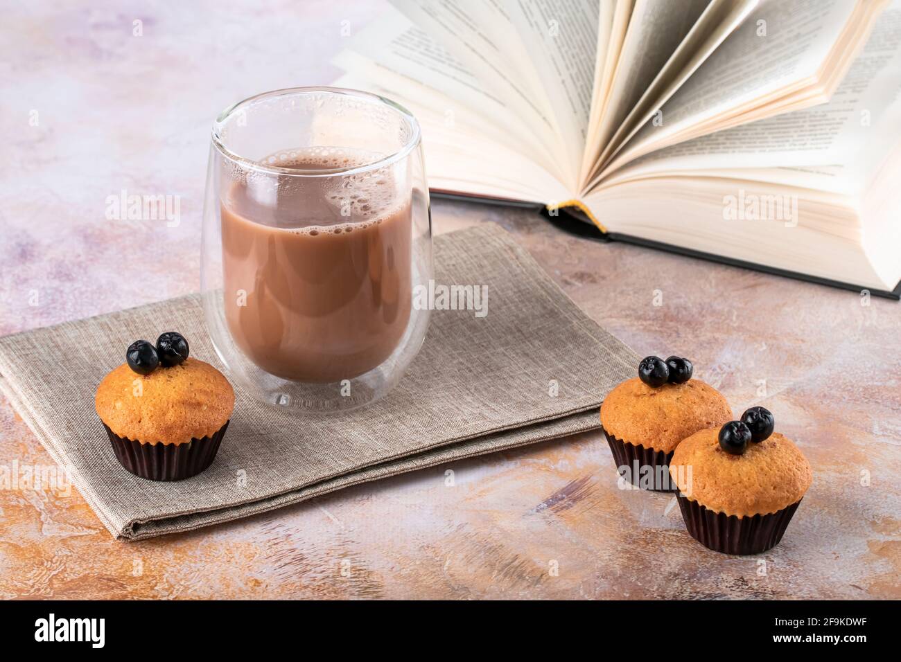 Muffin e un bicchiere di caffè bianco sul tavolo. Bevande calde, cacao e libro cartaceo aperto. Vita still rustica in stile vintage. Tortini marroni con frutti di bosco. Foto Stock
