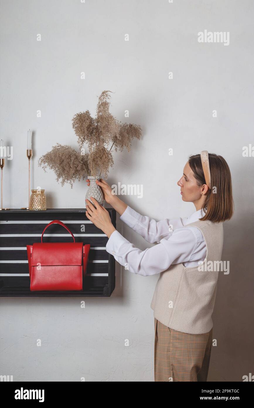 Donna stilista di moda millenario decora uno scaffale con un rosso borsa con vaso in erba di pampas Foto Stock