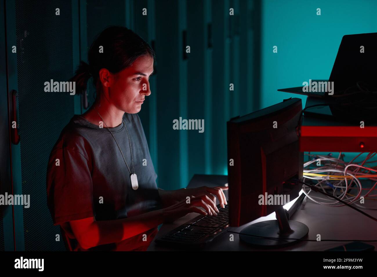 Vista laterale ritratto di un tecnico IT femminile che utilizza il computer mentre lavora in una sala server illuminata da luce rossa, spazio di copia Foto Stock
