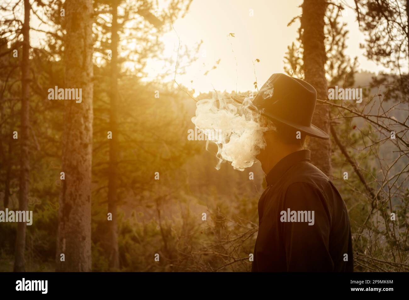 Cowboy che fuma nella foresta.Male in cappello nero e camicia scura con nuvola di fumo al tramonto.bello paesaggio creativo di bosco con spazio di copia. Foto Stock