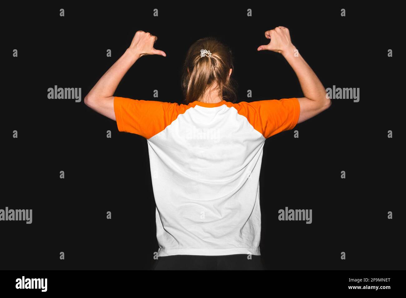 Una ragazza in una T-shirt su uno sfondo nero punta con le mani alla schiena, un luogo per il testo. Progettare i vestiti dello spazio di copia. Foto Stock