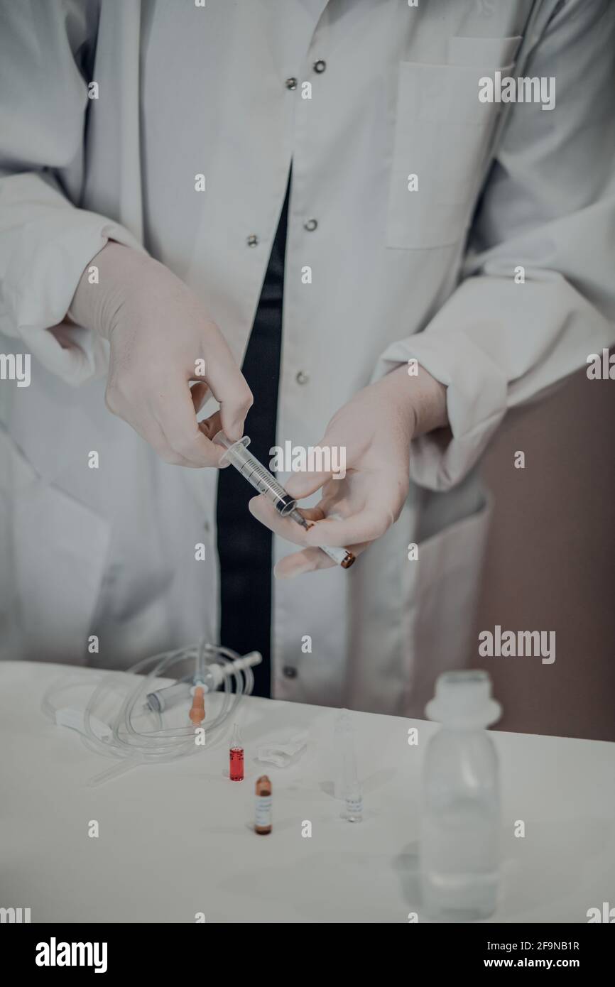 Mani femminili in guanti medici bianchi che disegnano il medicinale dalla fiala in una siringa, accanto alla fiala verticale con il medicinale su un tavolo acrilico bianco. Liquido Foto Stock