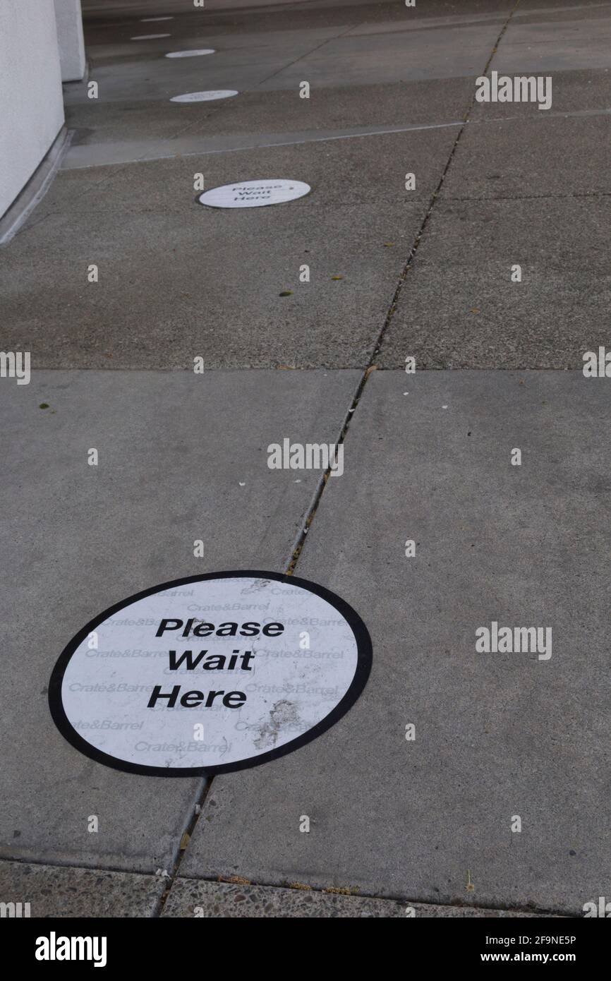 "Attendi qui" legge il cartello sul cemento all'esterno dell'ingresso di un grande magazzino, uno strumento di social distancing durante la pandemia COVID-19. Foto Stock