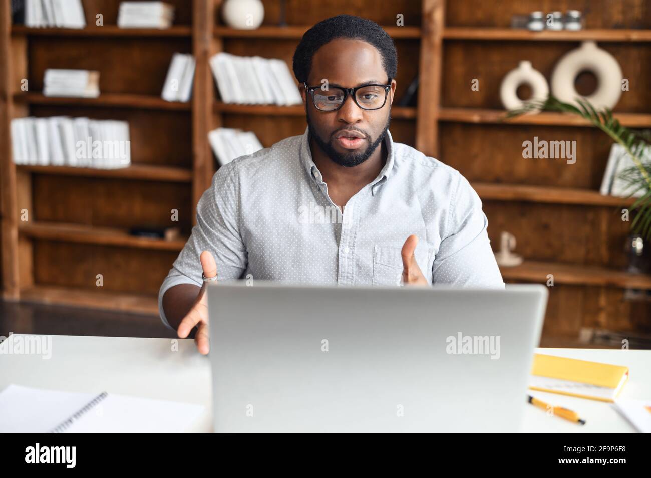 Concentrato giovane operaio afroamericano in occhiali guardando la webcam del laptop e gesturing, uomo nero sicuro utilizzando l'app compter per la connessione video con i colleghi Foto Stock