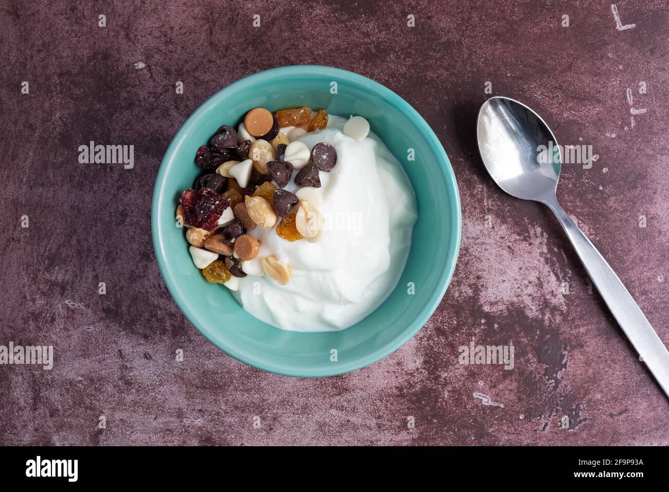 Vista dall'alto di una ciotola con yogurt greco puro e una miscela di pista di caramelle su un piano di marronte con un cucchiaio a lato. Foto Stock
