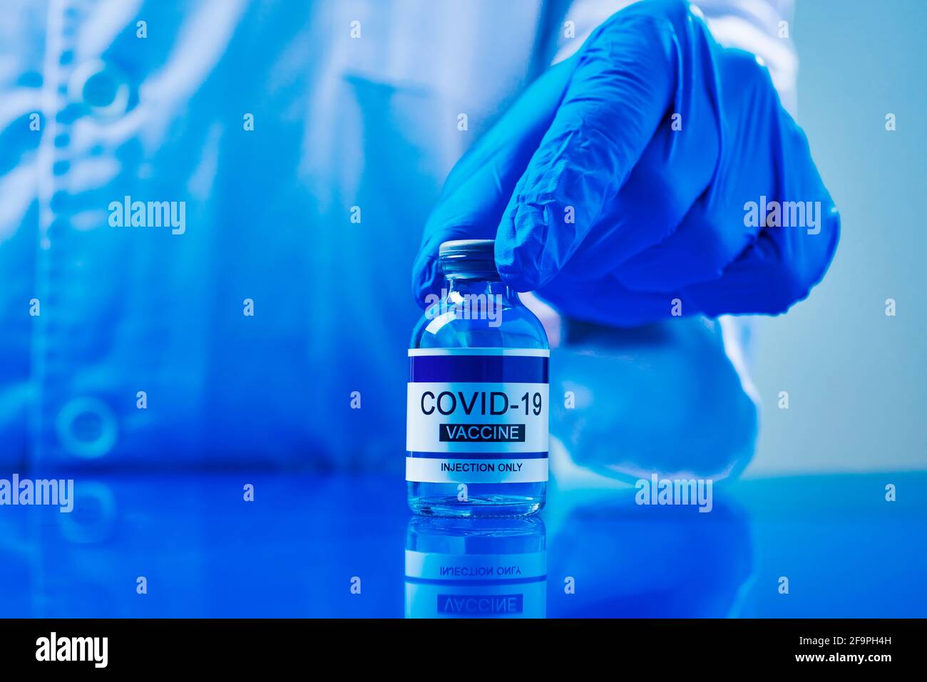 un operatore sanitario, indossando guanti chirurgici blu, mostra una bottiglia vaccinale di covid-19 simulata su un tavolo blu Foto Stock