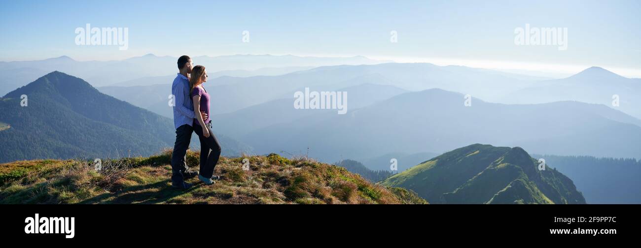 Tutta la lunghezza della coppia innamorata in piedi su una collina erbosa e guardando le belle montagne. Giovane viaggiatore abbracciando affascinante donna da dietro e ammirando la bellezza della natura. Vista panoramica Foto Stock