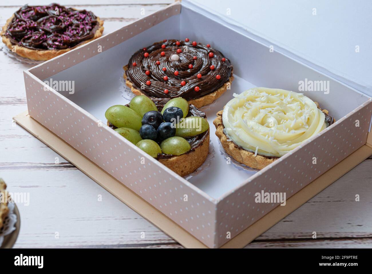Closeup di torta con pasta di burro farcito con ganache al cioccolato, decorato con uve verdi e mirtilli. All'interno di una confezione regalo e con altre torte. Foto Stock