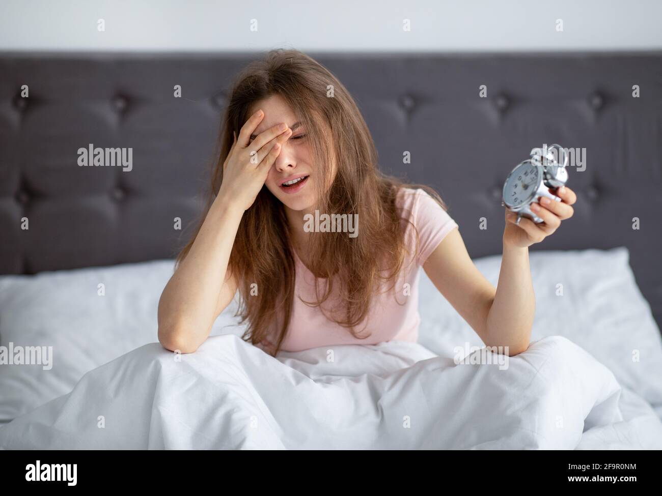 Giovane donna depressa con sveglia seduta a letto, piangendo in disperazione, non disposto a mettersi al lavoro, al chiuso Foto Stock