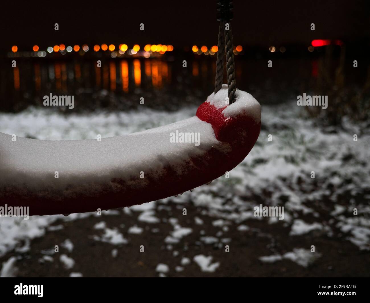 Altalena di corda di plastica rossa coperta da crosta di neve sulla riva del fiume in una serata invernale. Le luci notturne arancioni, rosse e gialle della città si riflettono nel fiume nero Foto Stock