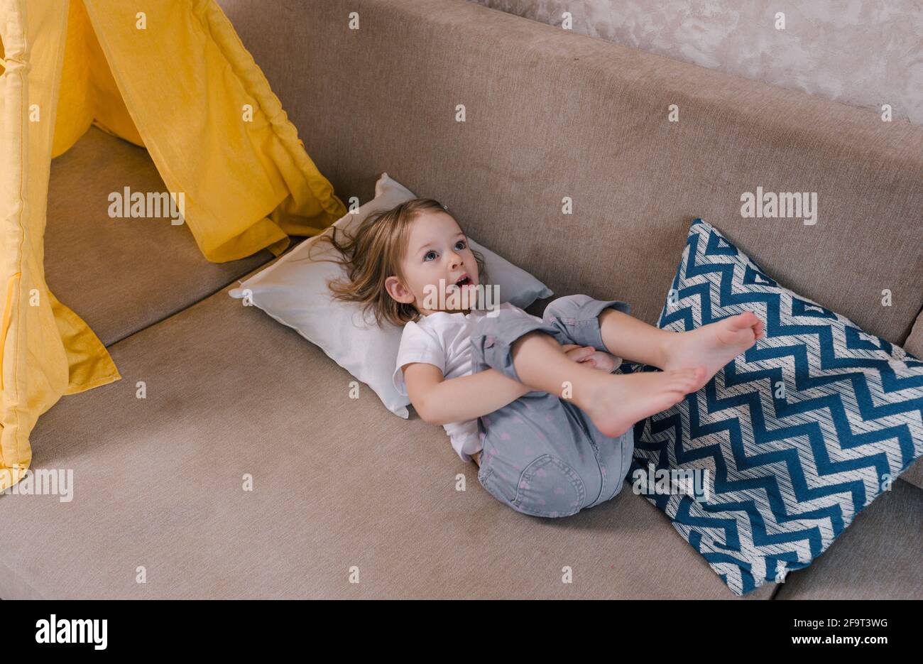 una bambina sta sdraiata sul divano sulla schiena con le gambe in su. giochi  per bambini a casa Foto stock - Alamy