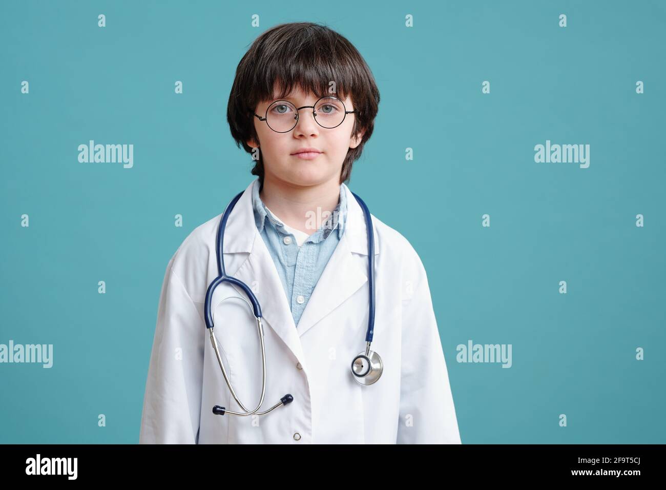Ritratto del ragazzino in camice bianco che gioca in medico guardando la fotocamera sullo sfondo blu Foto Stock